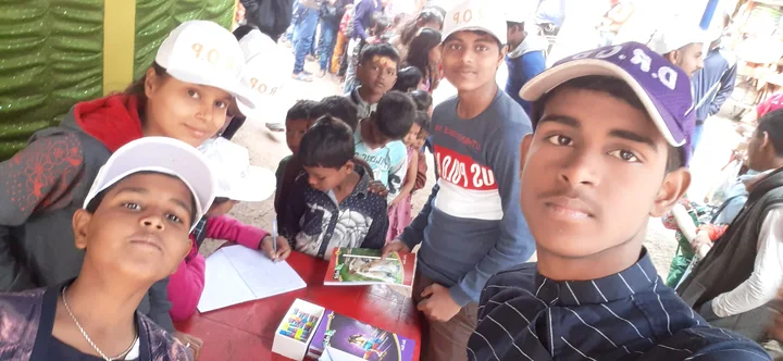 Children and volunteers in DROP caps gathered around books and a sign-in sheet during an education outreach activity.
