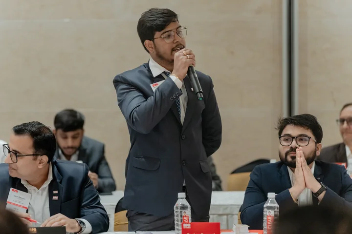 Roshan Burnwal speaking into a microphone at a BNI meeting, with fellow members seated nearby.