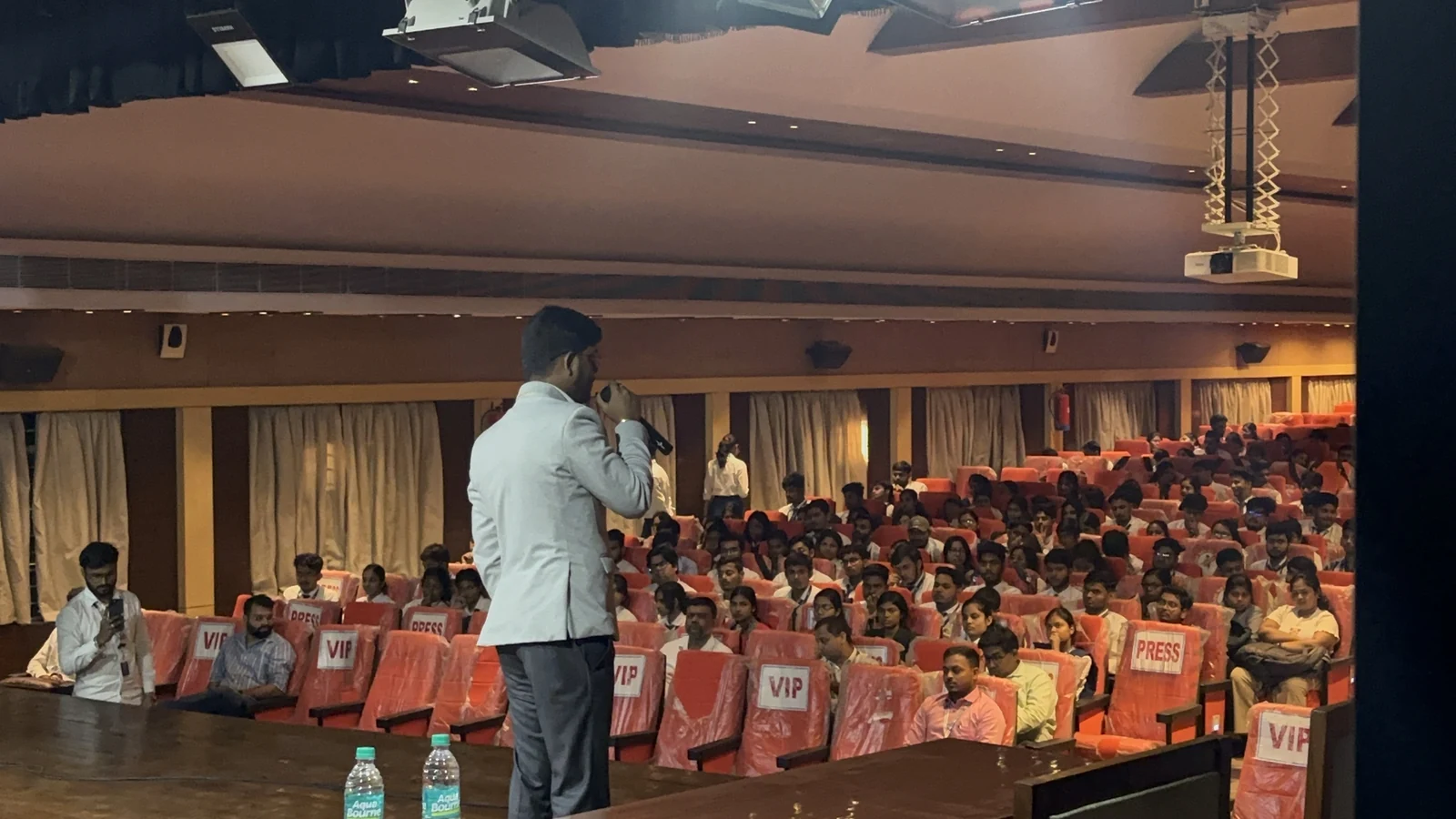 Roshan Burnwal speaking into a microphone to an auditorium audience at the Entrepreneurship Awareness Drive in Durgapur.